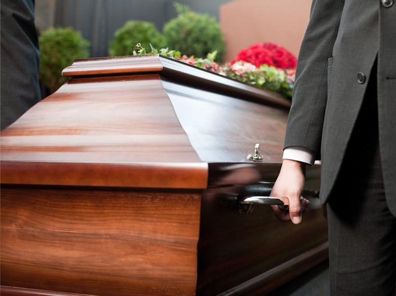 Eine Person in einem Anzug, die den Griff eines Holzsarges hält, welcher mit roten Rosen und Blumen geschmückt ist.